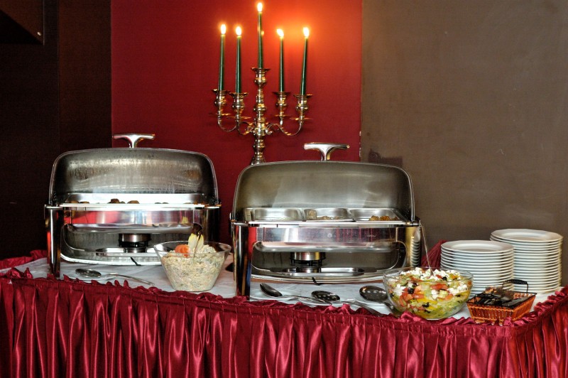 Dwa podgrzewacze bufetowe ze stali nierdzewnej, sałatki w szklanych misach, stosy białych talerzy i świecznik z zapalonymi świecami na stole z bordową falbaną.